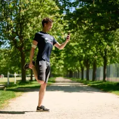 Un homme fait un étirement quadriceps sur un chemin en terre, regardant son téléphone.