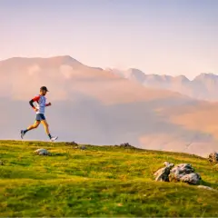 Un coureur en plein effort sur une crête herbeuse, avec des montagnes brumeuses en arrière-plan. Il participe au **championnat du monde de trail long**.