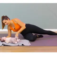 Une femme souriante fait du sport après accouchement, en position de planche latérale, avec son bébé allongé à côté sur un tapis.