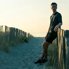 Homme sportif, en tenue de course, se repose contre une clôture de bois sur une plage au coucher du soleil. Il a atteint son poids de forme.