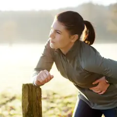 Une femme en tenue de sport s'appuie sur un poteau en bois, tenant son flanc droit, visiblement prise d'un **point de côté** lors de son entraînement matinal.