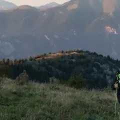 Un coureur de trail avec des bâtons monte une colline herbeuse, avec des montagnes en arrière-plan.