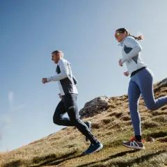 Couple en pleine préparation ultra trail 100 km, courant sur un sentier de montagne rocailleux sous un ciel bleu.