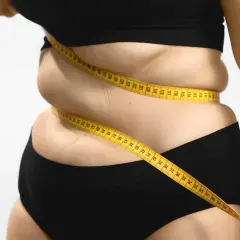 Une femme mesure son tour de taille avec un mètre ruban jaune, symbolisant le suivi du pourcentage masse grasse et le parcours de perte de poids.