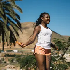 Femme souriante faisant de l'exercice corde à sauter sous un ciel bleu, entourée de palmiers et de collines.