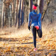 Une coureuse fait un étirement après trail dans une forêt automnale, entourée de feuilles mortes et d'arbres.
