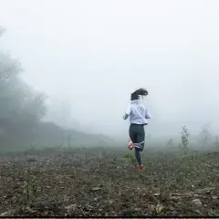 Femme courant dans le brouillard, une scène de préparation physique trail. Le paysage est brumeux, avec des arbres et un terrain rocailleux.