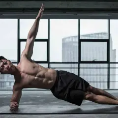 Un homme musclé réalise un exercice oblique, bras levé, dans une salle de sport avec vue sur la ville.