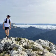 Un homme en tenue de trail, chapeau et lunettes, marche sur un sentier rocheux avec une vue imprenable sur les montagnes. Un week-end choc trail inoubliable.