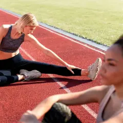 Deux femmes font un exercice de mobilité sur une piste d'athlétisme. L'une est assise, jambe tendue, l'autre est floue au premier plan.