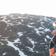 Femme souriante, en tenue de sport, assise sur des rochers en montagne, vérifiant sa montre. Préparation ultra trail dans un paysage enneigé.