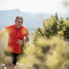 Un homme en tenue de sport orange et noire, souriant, prêt à commencer la course à pied sur un sentier de montagne.