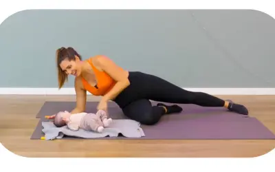 Une femme souriante fait du sport après accouchement, en position de planche latérale, avec son bébé allongé à côté sur un tapis.