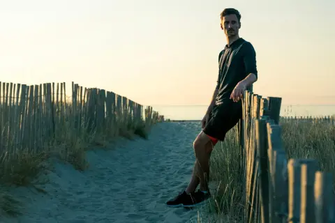 Homme sportif, en tenue de course, se repose contre une clôture de bois sur une plage au coucher du soleil. Il a atteint son poids de forme.