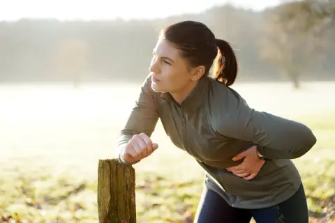 Une femme en tenue de sport s'appuie sur un poteau en bois, tenant son flanc droit, visiblement prise d'un **point de côté** lors de son entraînement matinal.