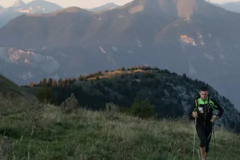 Un coureur de trail avec des bâtons monte une colline herbeuse, avec des montagnes en arrière-plan.