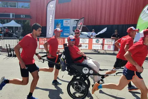 Une équipe court avec un homme en fauteuil roulant lors d'une course en joëlette. Ils portent tous des t-shirts rouges et des casquettes.