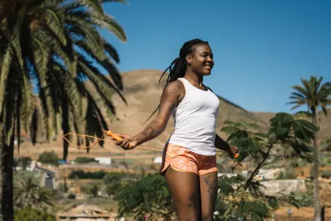 Femme souriante faisant de l'exercice corde à sauter sous un ciel bleu, entourée de palmiers et de collines.