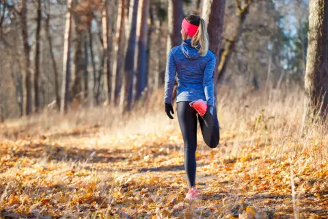 Une coureuse fait un étirement après trail dans une forêt automnale, entourée de feuilles mortes et d'arbres.