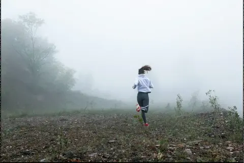 Femme courant dans le brouillard, une scène de préparation physique trail. Le paysage est brumeux, avec des arbres et un terrain rocailleux.