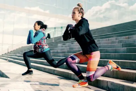 Deux femmes font des fentes sur des escaliers extérieurs, profitant d'une séance de ppg sport sous un ciel bleu.