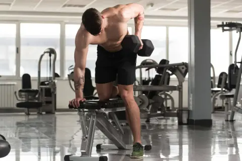 Un homme musclé effectue un exercice haltère, concentré sur son entraînement dans une salle de sport lumineuse.
