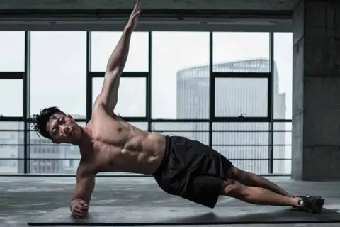 Un homme musclé réalise un exercice oblique, bras levé, dans une salle de sport avec vue sur la ville.