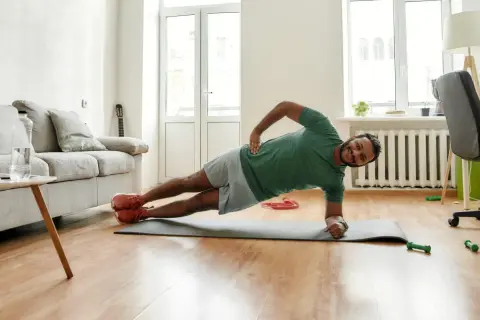 Un homme souriant pratique le gainage latéral sur un tapis de yoga dans son salon.