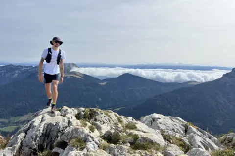 Un homme en tenue de trail, chapeau et lunettes, marche sur un sentier rocheux avec une vue imprenable sur les montagnes. Un week-end choc trail inoubliable.