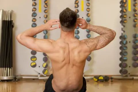 Homme musclé effectuant un exercice pour le dos, mains derrière la tête, dans une salle de sport avec des haltères en arrière-plan.