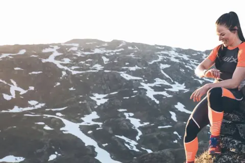 Femme souriante, en tenue de sport, assise sur des rochers en montagne, vérifiant sa montre. Préparation ultra trail dans un paysage enneigé.