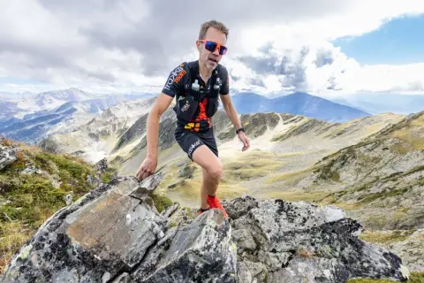 Un traileur en plein effort sur un sentier rocailleux, le regard déterminé, incarne l'ultra spirit dans un paysage montagneux spectaculaire.