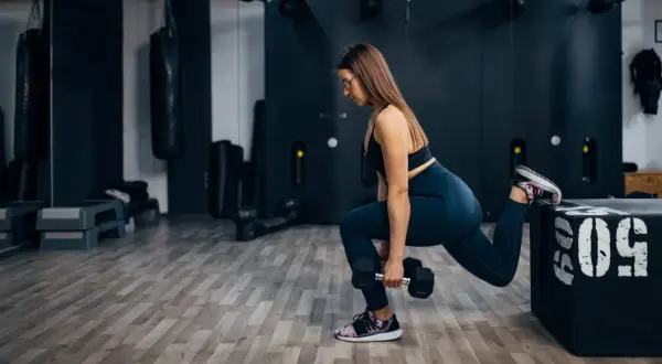 Femme effectuant une fente bulgare avec haltère, entraînement de jambes intense dans une salle de sport moderne.