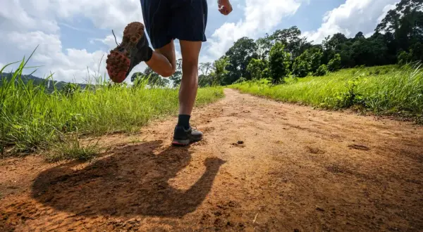 Un coureur s'élance sur un chemin de terre bordé d'herbes hautes, prêt pour un trail longue distance sous un ciel nuageux.