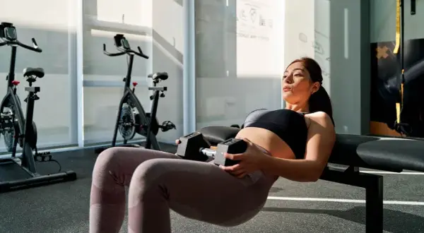Femme enceinte effectuant un hip thrust au sol avec haltères, concentrée sur son entraînement dans une salle de sport lumineuse.