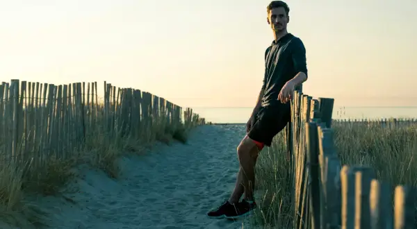 Homme sportif, en tenue de course, se repose contre une clôture de bois sur une plage au coucher du soleil. Il a atteint son poids de forme.