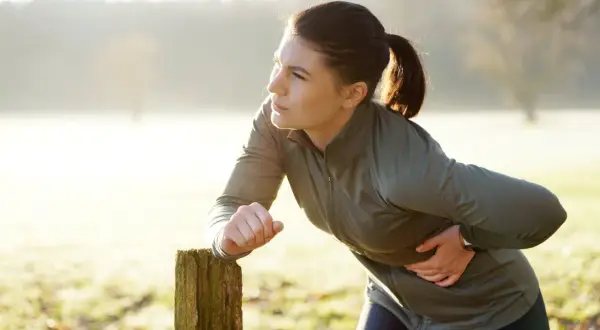 Une femme en tenue de sport s'appuie sur un poteau en bois, tenant son flanc droit, visiblement prise d'un **point de côté** lors de son entraînement matinal.