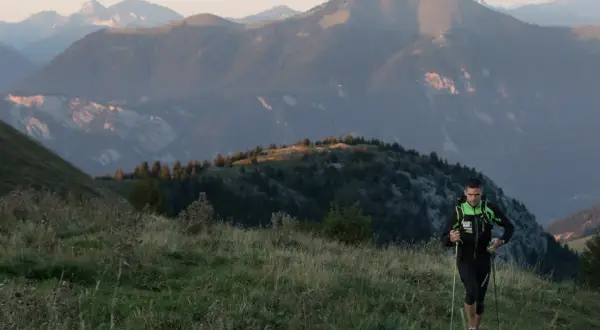 Un coureur de trail avec des bâtons monte une colline herbeuse, avec des montagnes en arrière-plan.