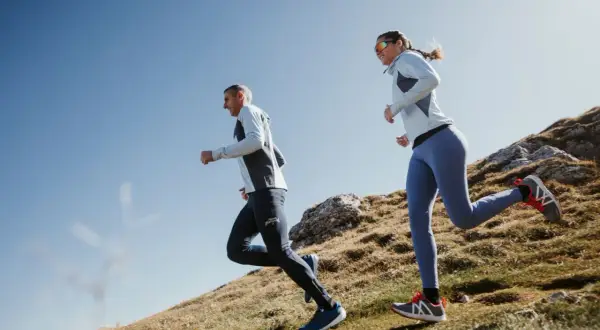Couple en pleine préparation ultra trail 100 km, courant sur un sentier de montagne rocailleux sous un ciel bleu.