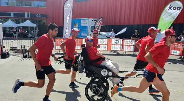 Une équipe court avec un homme en fauteuil roulant lors d'une course en joëlette. Ils portent tous des t-shirts rouges et des casquettes.