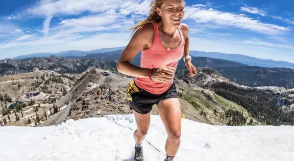Une coureuse en plein effort, le visage rayonnant, dévale une pente enneigée en montagne. Son **endurance active** est mise à l'épreuve dans ce paysage spectaculaire.