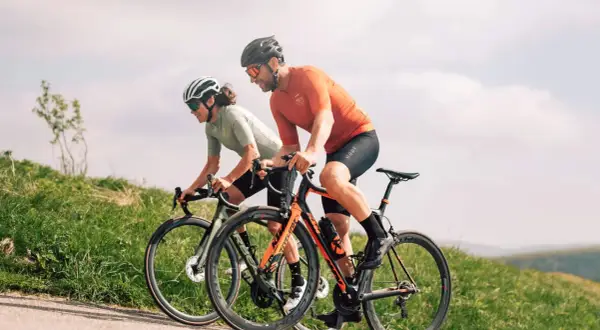 Deux cyclistes en plein **entrainement croisé** sur une route de campagne, profitant du paysage verdoyant sous un ciel nuageux.