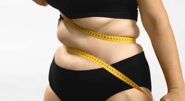 Une femme mesure son tour de taille avec un mètre ruban jaune, symbolisant le suivi du pourcentage masse grasse et le parcours de perte de poids.