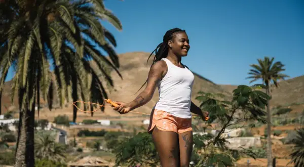 Femme souriante faisant de l'exercice corde à sauter sous un ciel bleu, entourée de palmiers et de collines.