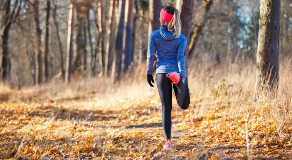 Une coureuse fait un étirement après trail dans une forêt automnale, entourée de feuilles mortes et d'arbres.