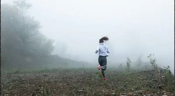 Femme courant dans le brouillard, une scène de préparation physique trail. Le paysage est brumeux, avec des arbres et un terrain rocailleux.