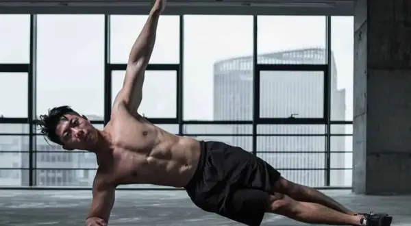 Un homme musclé réalise un exercice oblique, bras levé, dans une salle de sport avec vue sur la ville.