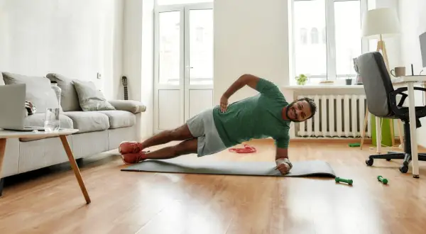 Un homme souriant pratique le gainage latéral sur un tapis de yoga dans son salon.