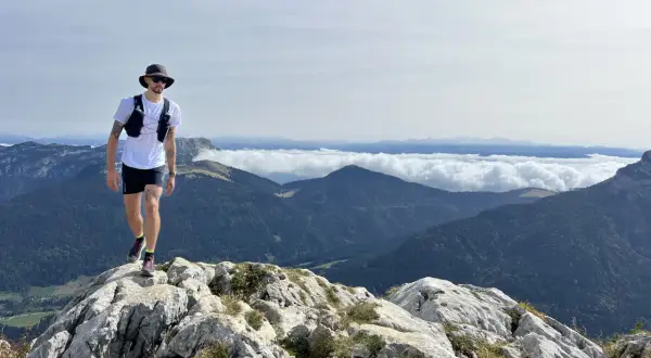 Un homme en tenue de trail, chapeau et lunettes, marche sur un sentier rocheux avec une vue imprenable sur les montagnes. Un week-end choc trail inoubliable.