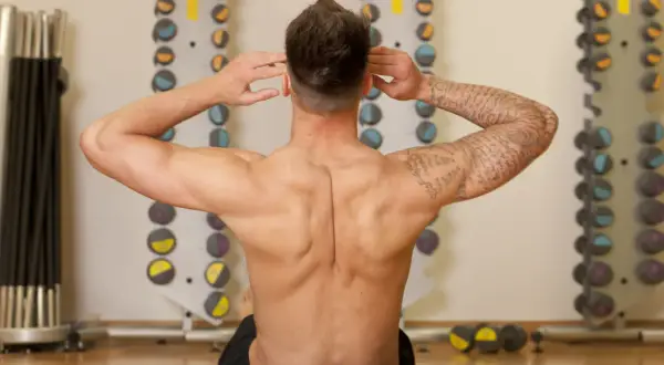 Homme musclé effectuant un exercice pour le dos, mains derrière la tête, dans une salle de sport avec des haltères en arrière-plan.